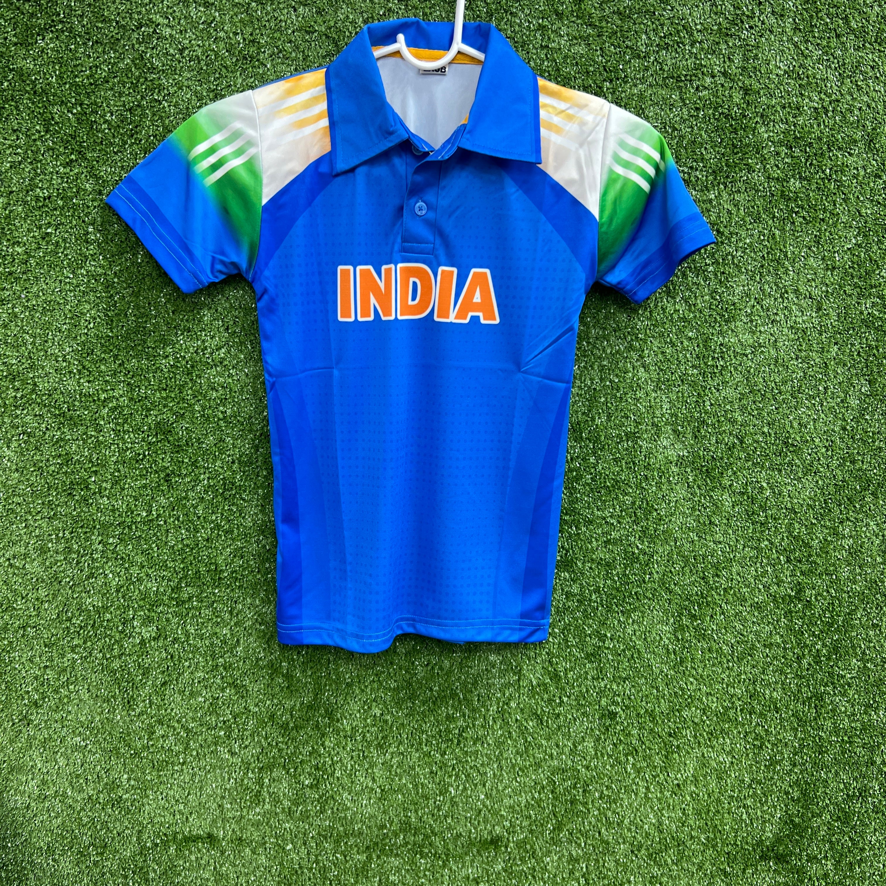 A full shot of the blue jersey laid flat, showing the orange "INDIA" text, collar, and tri-color sleeve details on a green grass background.