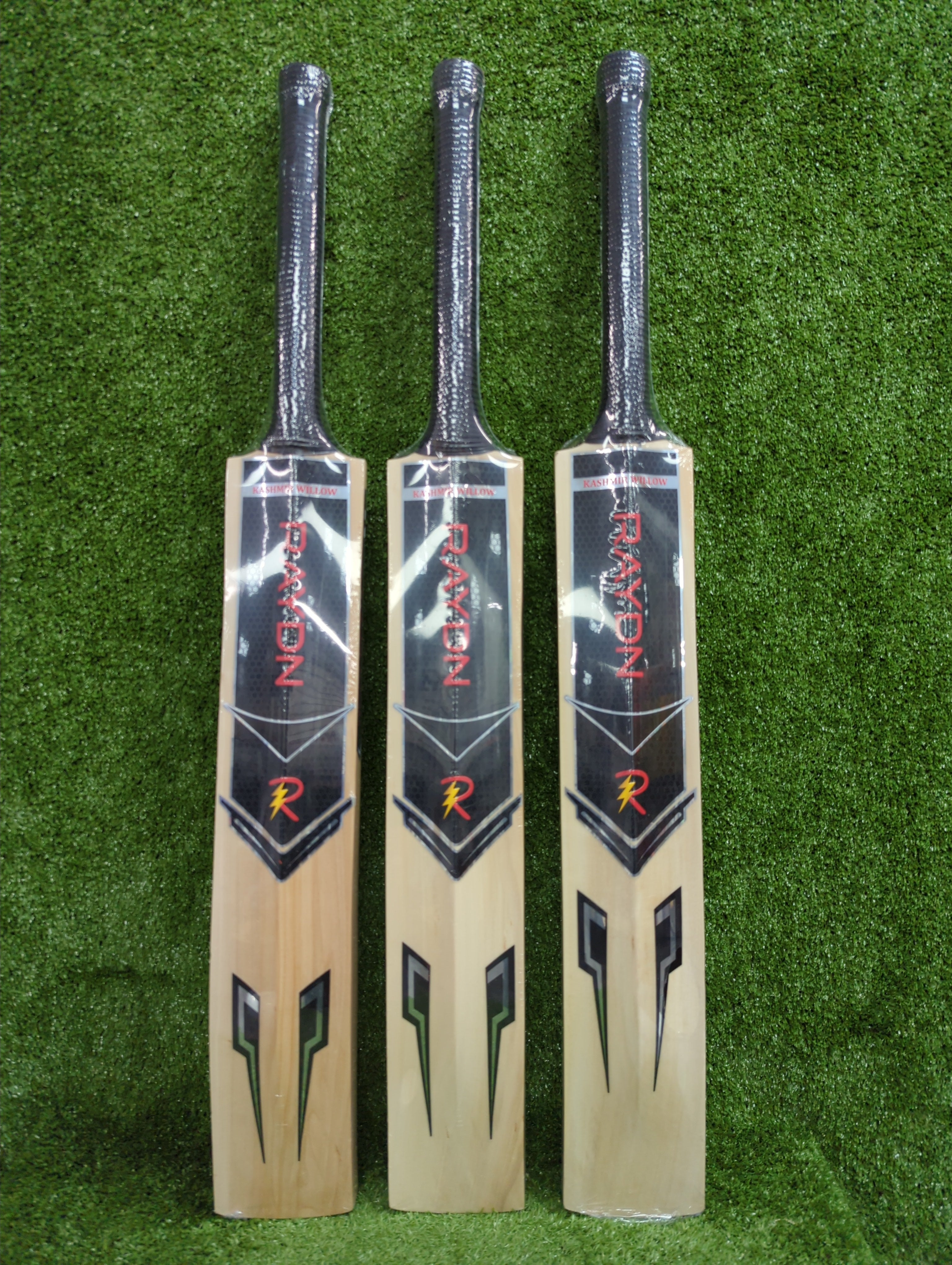 Back view of three Raydn VK Profile Kashmir Willow tennis cricket bats showing the full spine design and black, silver, and neon green branding on green grass.